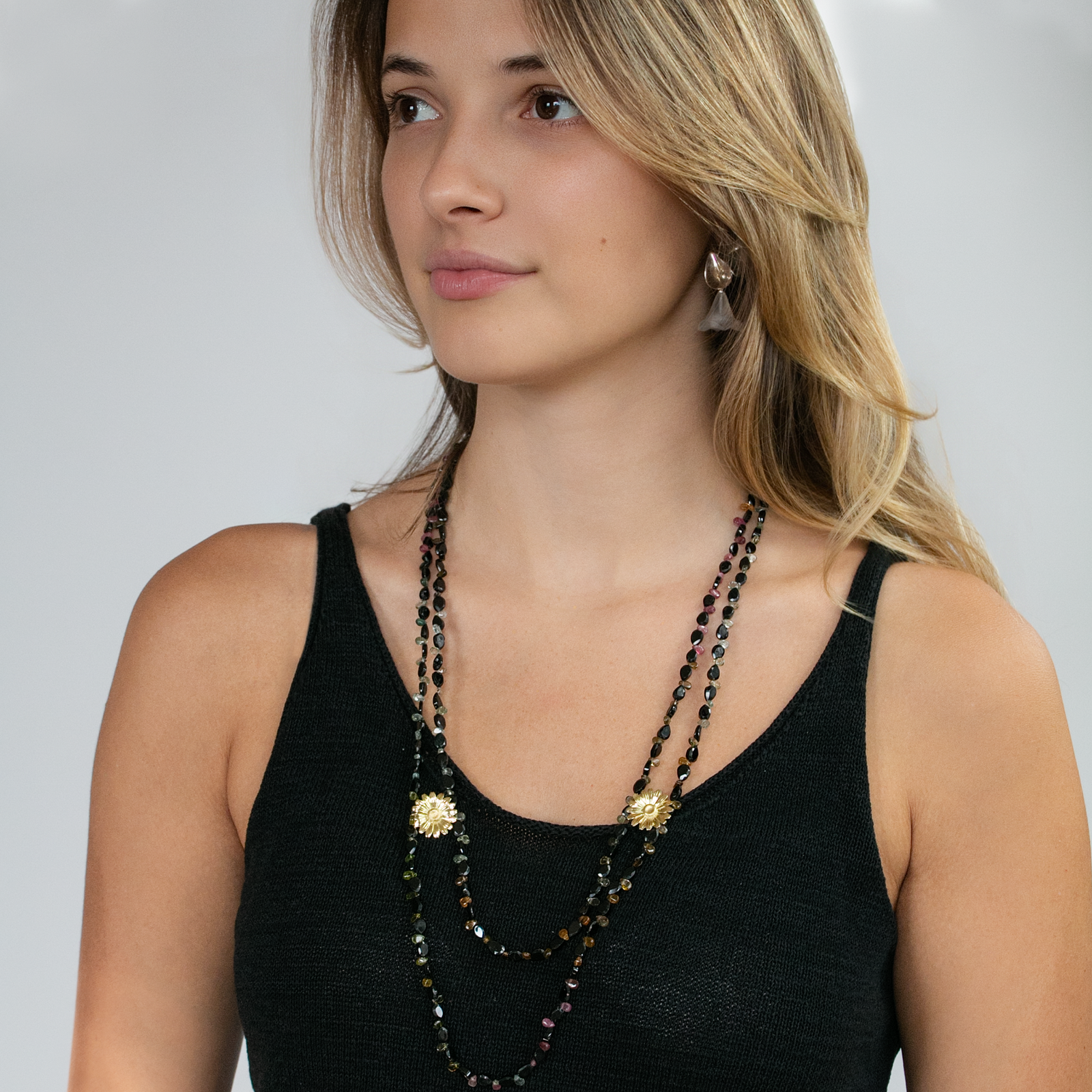 A woman wearing a double-strand beaded necklace with gold floral accents and dark iridescent beads, paired with a black tank top against a light gray background.